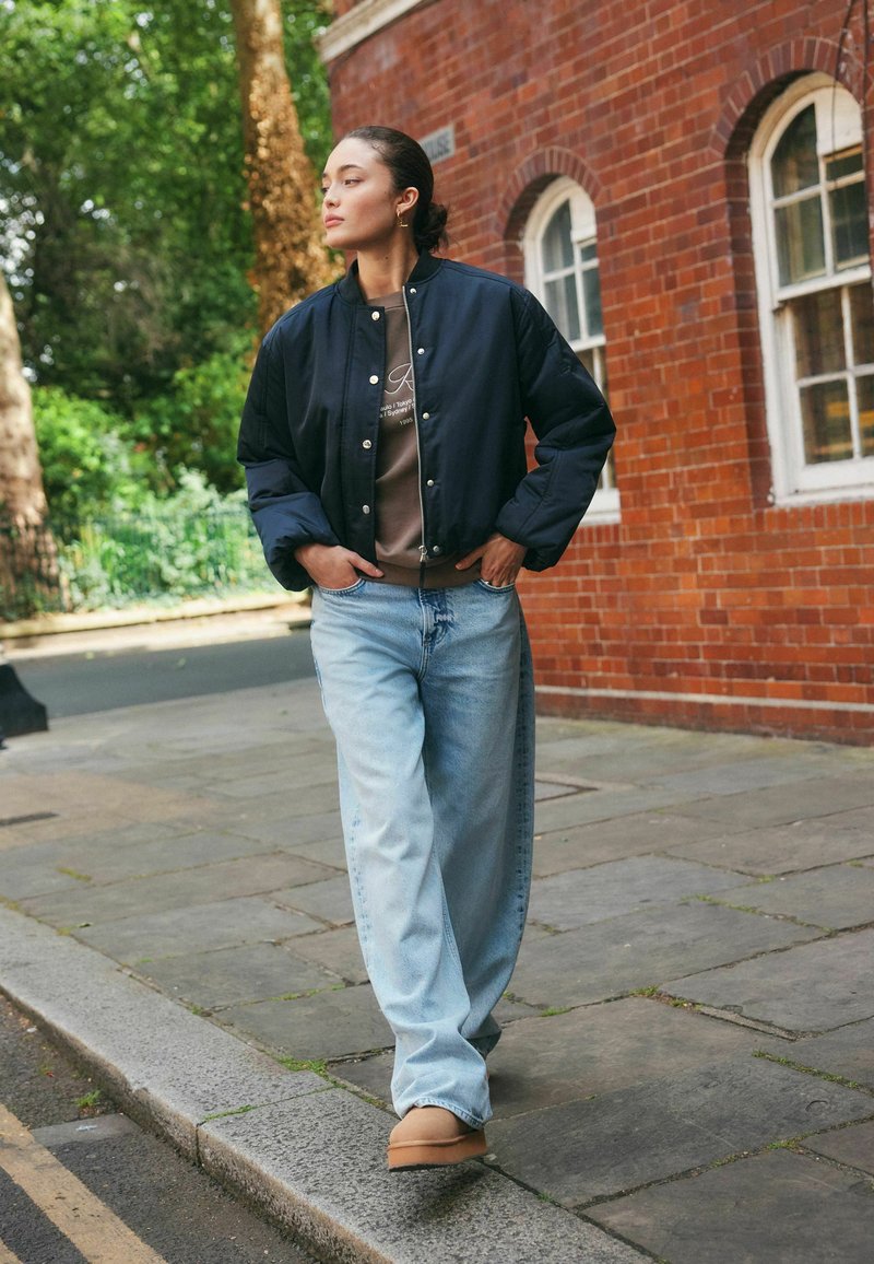 Junge Frau, die auf dem Bürgersteig neben einem Gebäude aus rotem Backstein geht, trägt eine marineblaue Bomberjacke, einen braunen Pullover, hellblaue Jeans und beige Schuhe.