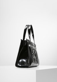Svart lackläder tote-väska med en glänsande yta, som har dubbla handtag och en cirkulär logotypdetalj på ena sidan. Sned vy.