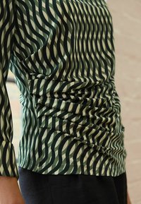 Top a righe verde e crema con maniche lunghe, caratterizzato da un motivo a onde e una texture arricciata sul lato. Abbina a pantaloni scuri.