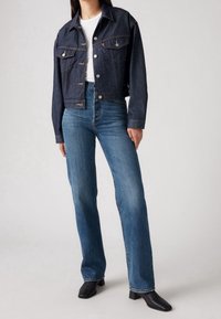 Chaqueta de mezclilla oscura cortada con cierre de botón, dos bolsillos frontales, combinada con vaqueros azul de talle alto y botas negras de punta cuadrada.