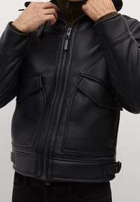 Giubbotto in pelle nera con zip frontale, due ampie tasche anteriori e dettagli a coste sulle spalle. La texture appare liscia con una leggera lucentezza.