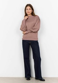 Pull léger mauve avec des manches bouffantes, des poignets côtelés et un ourlet côtelé. Associé à un jean bleu foncé évasé et des chaussures noires. Debout contre un mur blanc.