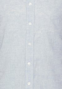 Jasnoniebieska koszula zapinana na guziki z teksturowanej tkaniny, z białymi guzikami i klasycznym kołnierzem. Szwy są widoczne wzdłuż listwy zapinającej.