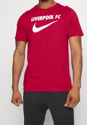 Mężczyzna w czerwonej koszulce z białym tekstem "LIVERPOOL FC" i białym logo Nike swoosh na klatce piersiowej, zestawionej z czarnymi spodniami.