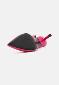 Une chaussure rose à talon haut posée sur le côté, montrant sa semelle noire pointue et son talon aiguille fin sur fond blanc.