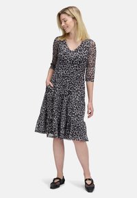 Frau in einem knielangen Leopardenmusterkleid mit durchsichtigen Dreiviertelärmeln, lächelnd und stehend mit einer Hand in der Tasche und schwarzen Schuhen.