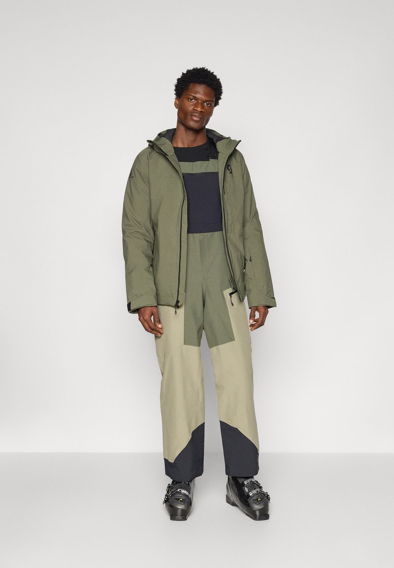 Izolowana kurtka w oliwkowym kolorze z kapturem, zestawiona z teksturowanym kombinezonem w dwóch odcieniach z łatami w jasnej zieleni i ciemnym niebieskim.