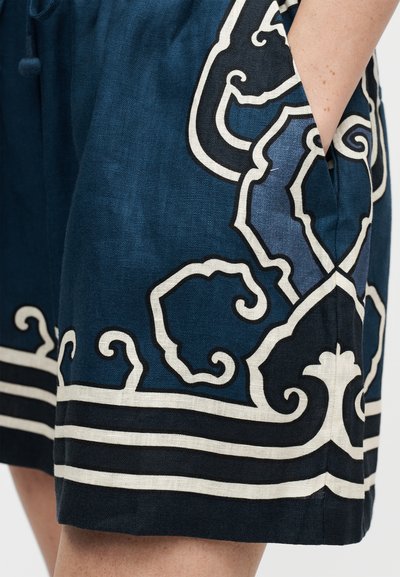 Μπλε σορτς με λευκά και μαύρα διακοσμητικά σπειροειδή σχέδια, που δείχνουν ένα χέρι μερικώς στην πλαϊνή τσέπη σε ανοιχτόχρωμο δέρμα.