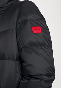 Chaqueta acolchada negra con diseño guateado, textura suave y una etiqueta rectangular roja con la palabra "HUGO" en la manga izquierda.