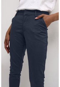 Gros plan sur une personne portant un pantalon bleu foncé avec une main dans la poche et une chemise blanche rentrée.