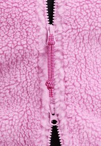 Rosa fleecejacka med texturerad yta, med en central dragkedja och flätad rosa dragrem. Dragkedjans tänder är svarta för ökad synlighet.