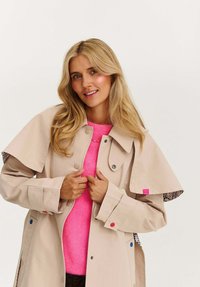 Blondýnka se usmívá, má na sobě béžový trenčkot přes jasně růžový svetr, drží klopy kabátu proti jednobarevnému pozadí.