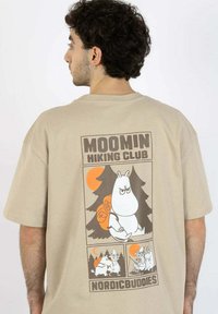 Valkoinen puuvillapaita, jossa on Moomin-hahmo takana teksteillä "Moomin Hiking Club" ja värikkäät sarjakuvakuvastot.