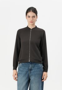 Svart zip-up jacka med ståkrage och långa ärmar. Slät textur, metallblixtlås, elastiska ärmslut och midja. Kombinerad med blå jeans.