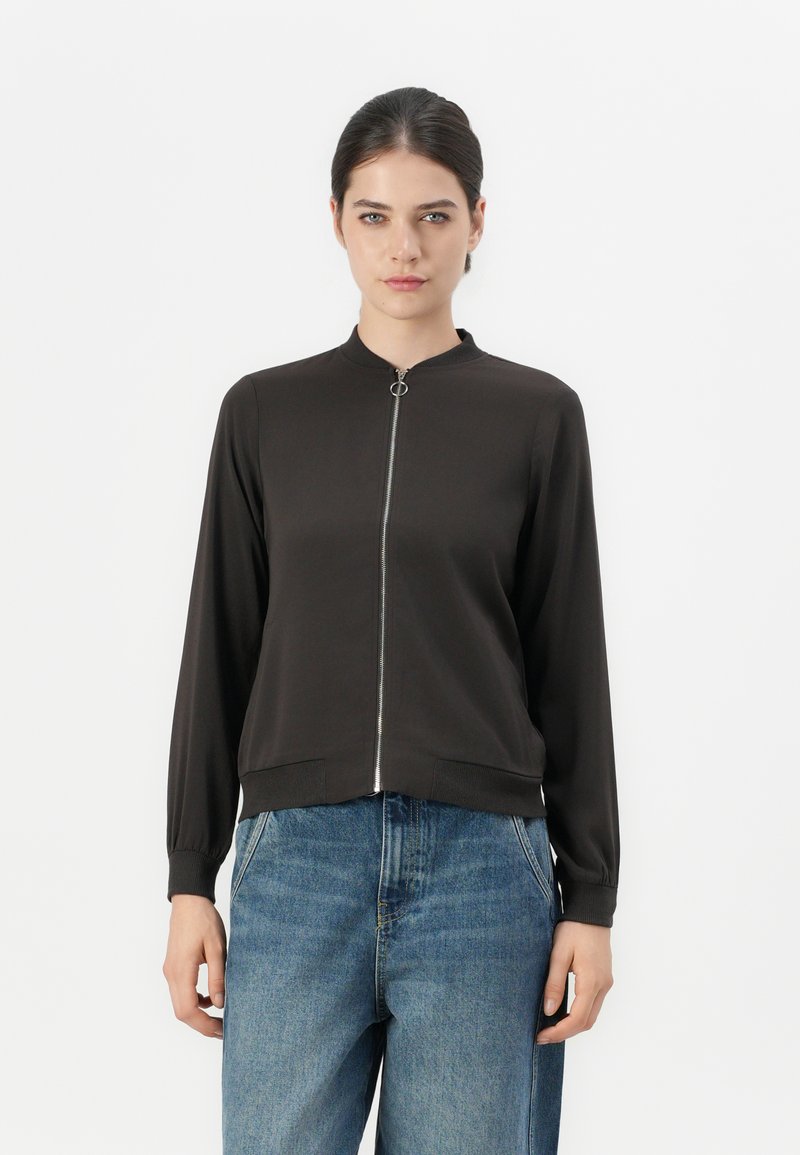 Svart zip-up jacka med ståkrage och långa ärmar. Slät textur, metallblixtlås, elastiska ärmslut och midja. Kombinerad med blå jeans.