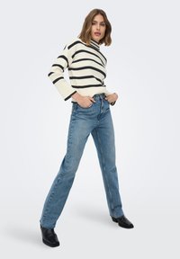 Gestreepte zwart-witte trui met een hoge halslijn, gecombineerd met lichtblauwe jeans met rechte pijpen en zwarte enkellaarzen.