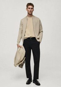 Beige pletený cardigan přes béžový svetr, černé kalhoty a černé boty. Drží světle béžovou bundu. Jednoduchý, moderní design s neutrálními barvami.