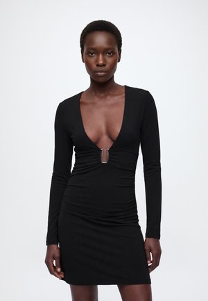 Mujer con un vestido mini negro ajustado de manga larga con un escote en V profundo y un detalle central de hebilla plateada, de pie frente a un fondo liso.