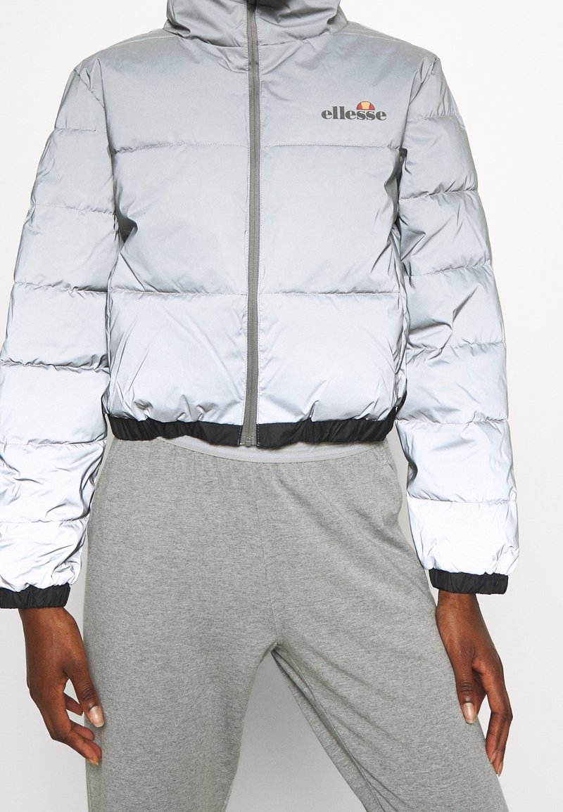 Personne portant une doudoune courte gris clair avec le logo ellesse et un pantalon de survêtement gris, debout devant un fond uni.