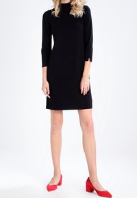 Robe noire longueur genou avec des manches trois-quarts, un col rond et une coupe ajustée. Associée à des escarpins rouges à bout pointu.