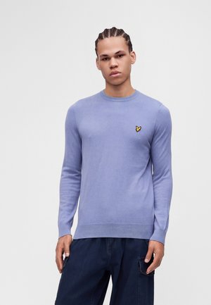 Jeune homme aux cheveux tressés portant un pull bleu clair à manches longues et un pantalon bleu foncé sur fond blanc uni.