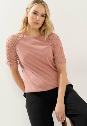 Vrouw met blond haar, gekleed in een stoffig roze top met korte kanten mouwen en zwarte broek, zittend tegen een effen lichte achtergrond.