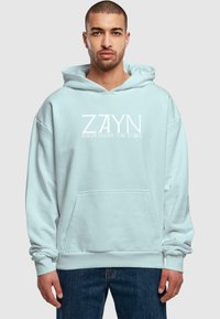 Lyseblå hættetrøje med en front kangaroo-lomme, snørehætte og hvid trykt tekst: "ZAYN ROOM UNDER THE STAIRS." Blødt materiale.