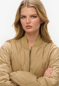 Gequilted tan bomberjack met ritssluiting en ribbelkraag. Beschikt over oversized mouwen en een textuur met golvenpatroon door het hele jack.