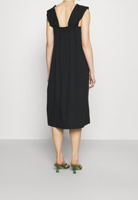 Černé bezramínkové šaty s volánovým designem na zadní straně, délka do poloviny lýtek, vyrobené z jemné tkaniny. Nosené s zelenými nazouvacími sandály na podpatku.