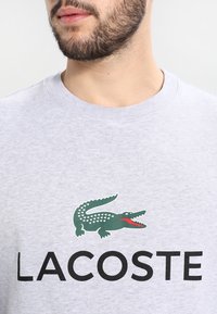 Felpa grigia in cotone con un logo di un coccodrillo verde con accenti rossi sopra la scritta "LACOSTE" in lettere nere grassetto.