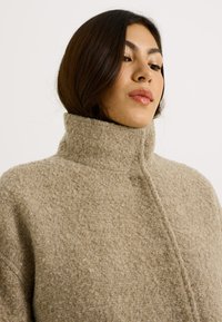 Mujer joven con cabello oscuro, vestida con un suéter de lana beige texturizado y de cuello alto, mirando ligeramente hacia abajo a su izquierda.