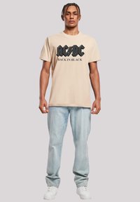 Beige cotton t-shirt featuring a black AC/DC logo and "BACK IN BLACK" text, paired with light blue denim jeans and white sneakers.
