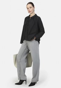 Zwarte blouse met kraag en strikdetail, gecombineerd met grijze wijde jeans. Accessoires omvatten een grote lichtgrijze tote en zwarte hoge hakken.