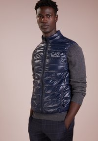 Gilet imbottito blu navy con design trapuntato, chiusura a zip e logo EA7, indossato sopra un maglione grigio. Caratterizzato dalla sua texture lucida e dalla forma aderente.