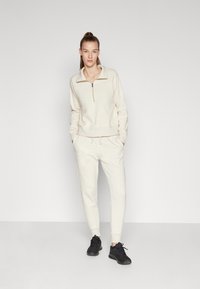Set de hanorac și pantaloni din fleece bej deschis. Guler cu fermoar, manșete și talie ribbate. Accente contrastante cu logo, design slim-fit. Adidași negri.