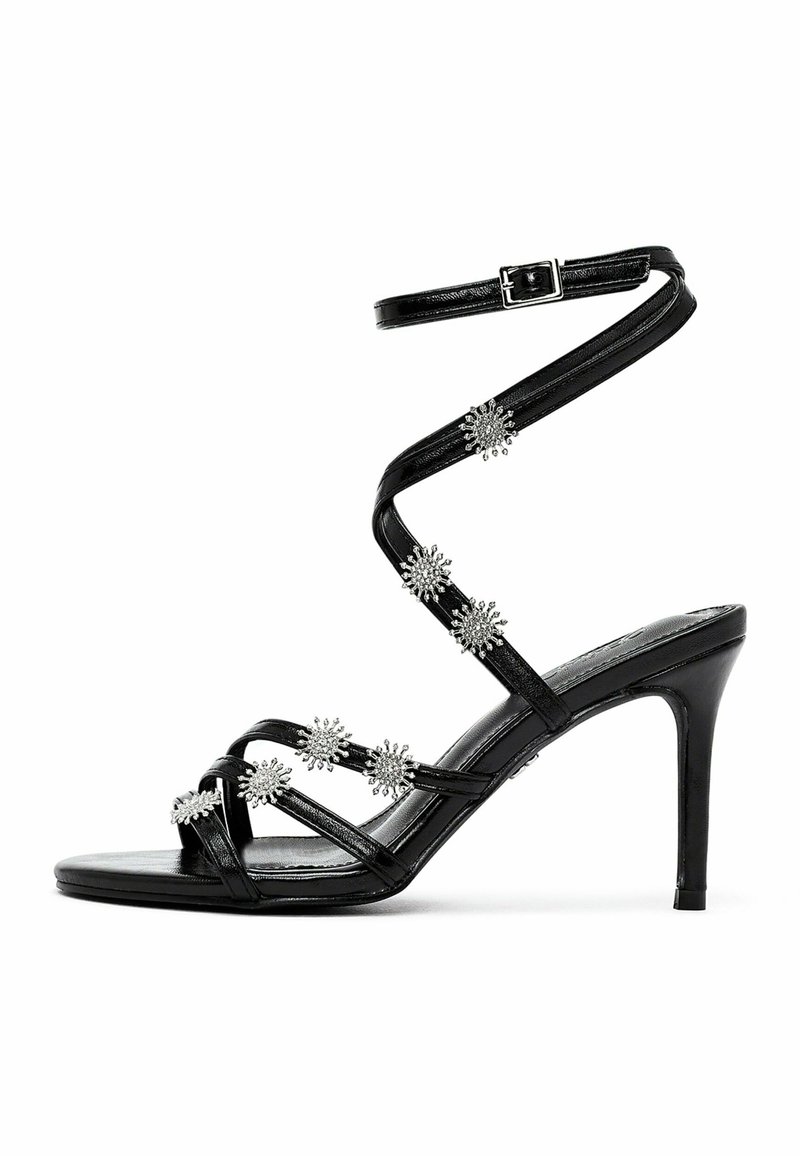 Sandale à talons hauts noire à lanières avec des accents métalliques et une sangle de cheville à boucle, comportant des embellissements floraux le long des lanières.