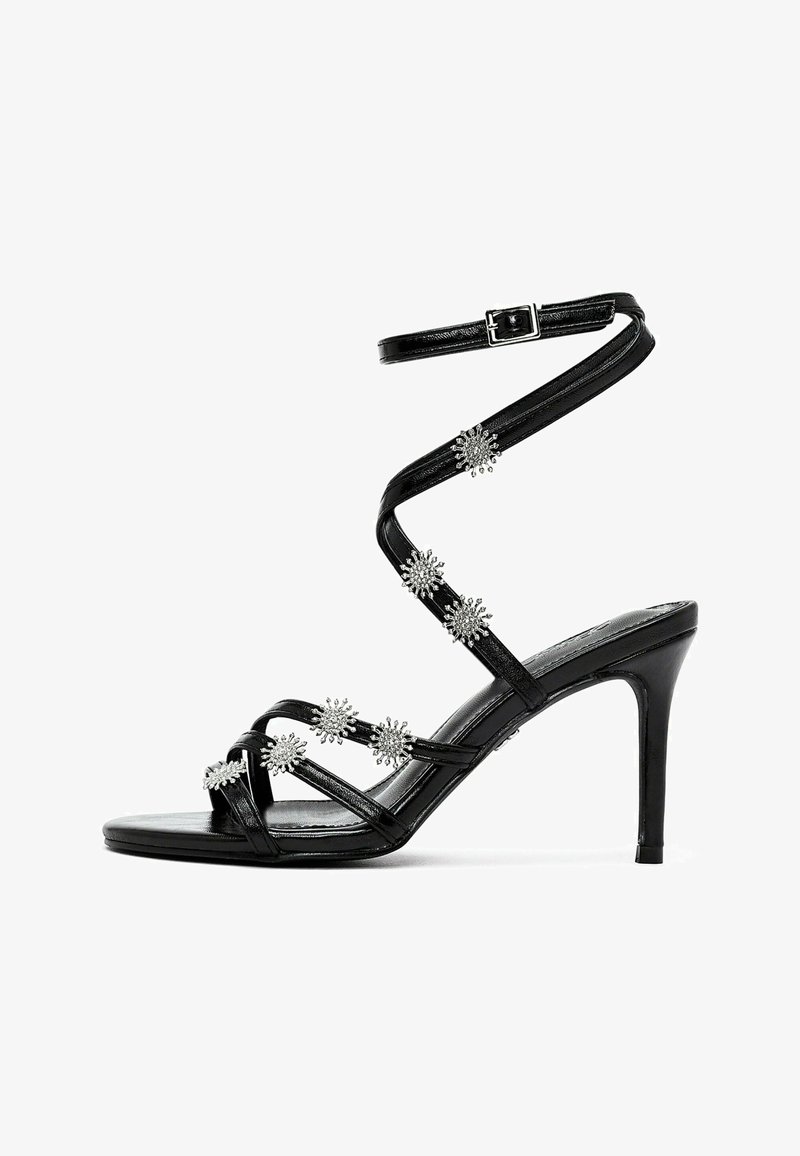 Sandale à talons hauts noire à lanières avec des accents métalliques et une sangle de cheville à boucle, comportant des embellissements floraux le long des lanières.