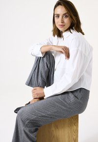 Witte button-down shirt met een relaxte fit, gecombineerd met grijze broek met visgraatpatroon. Model zit op een houten blok met zwarte instappers.