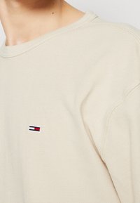 Beżowa, teksturowana koszula z długim rękawem i okrągłym dekoltem. Posiada małe, haftowane logo w kolorach czerwonym, białym i niebieskim na piersi.