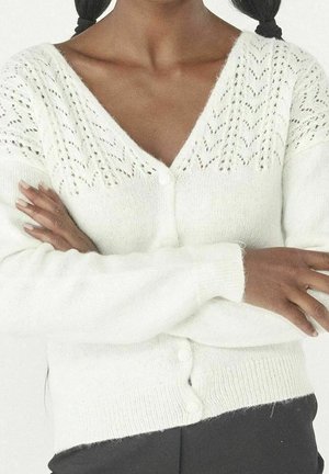 Frau mit mittel-dunkler Hautfarbe, die eine weiße Strickjacke mit Puffärmeln und Knöpfen trägt, die Arme verschränkt, und schwarze Hosen.