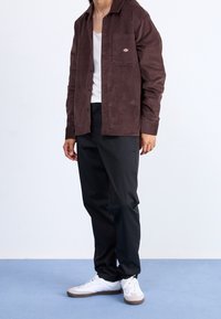 Chemise boutonnée en velours côtelé marron avec poche avant, associée à un pantalon noir et des baskets blanches avec des accents gris. Texture lisse, coupe décontractée.