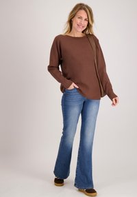 Bruine ongestructureerde trui met brede mouwen, gecombineerd met medium-wash wijde jeans en bruine instapschoenen. Neutrale achtergrond benadrukt de outfit.