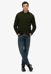 Sudadera de un verde oscuro con cuello simulado, presenta un bolsillo en el pecho. Combinada con jeans azules y zapatos negros, mostrando un ajuste relajado.