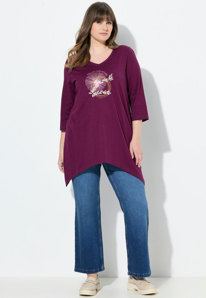 Lila V-Ausschnitt-Tunika mit Dreiviertelärmeln, asymmetrischem Saum und floralem Grafikdesign. Kombiniert mit weit geschnittenen blauen Denim-Jeans.