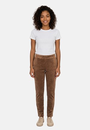 Donna con i capelli ricci che sorride, indossa una camicia bianca a maniche corte, pantaloni marroni e scarpe da ginnastica beige, in piedi davanti a uno sfondo semplice.