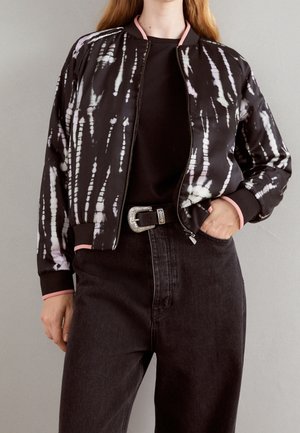 Person trägt eine schwarz-weiß batik Bomberjacke mit pinkem Besatz, schwarzes Top, dunkle Jeans und einen Gürtel mit großer silberner Schnalle.