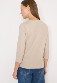 Haut beige à manches longues avec un col rond, une texture lisse et une coupe ajustée, associé à un jean en denim bleu, vu de dos.
