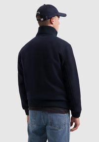 Donkerblauwe bomberjack met een geribbelde kraag en manchetten. Draagt over een gestreept shirt met een textuur stof en een ontspannen pasvorm.