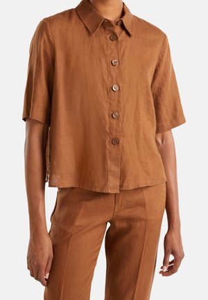 Femme portant une chemise marron à manches courtes boutonnée et un pantalon taille haute assorti, sur un fond blanc.