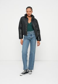 Černá péřová bunda s kratším střihem, nošená přes zelený top. Světle modré džíny rovného střihu a černé vysoké tenisky dotvářejí celý outfit.